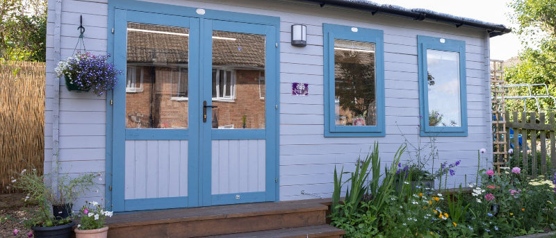 The Rowan Room. Log Cabin surrounded by flowers with blue door and windows.